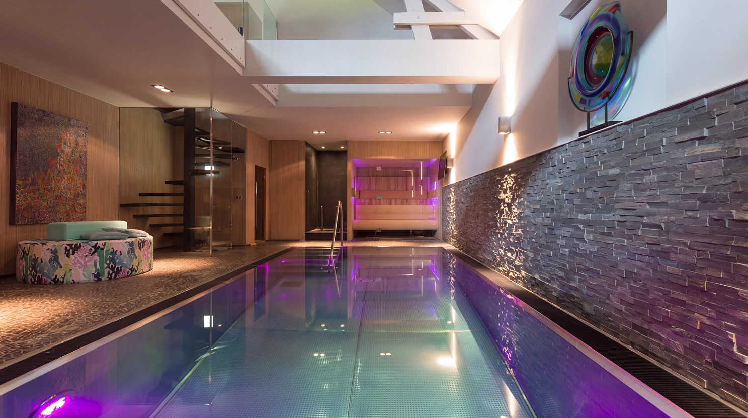 Intérieur d'un showroom moderne et lumineux exposant des modèles de piscines et équipements wellness. Au premier plan, une petite piscine de démonstration aux parois vitrées montrant l'eau turquoise. Derrière, un sauna en bois clair et un spa jacuzzi blanc sont visibles sur des socles de présentation. Des brochures et catalogues sont disposés sur un comptoir d'accueil. Éclairage professionnel, sol en carrelage gris clair, atmosphère accueillante et moderne mettant en valeur les installations.