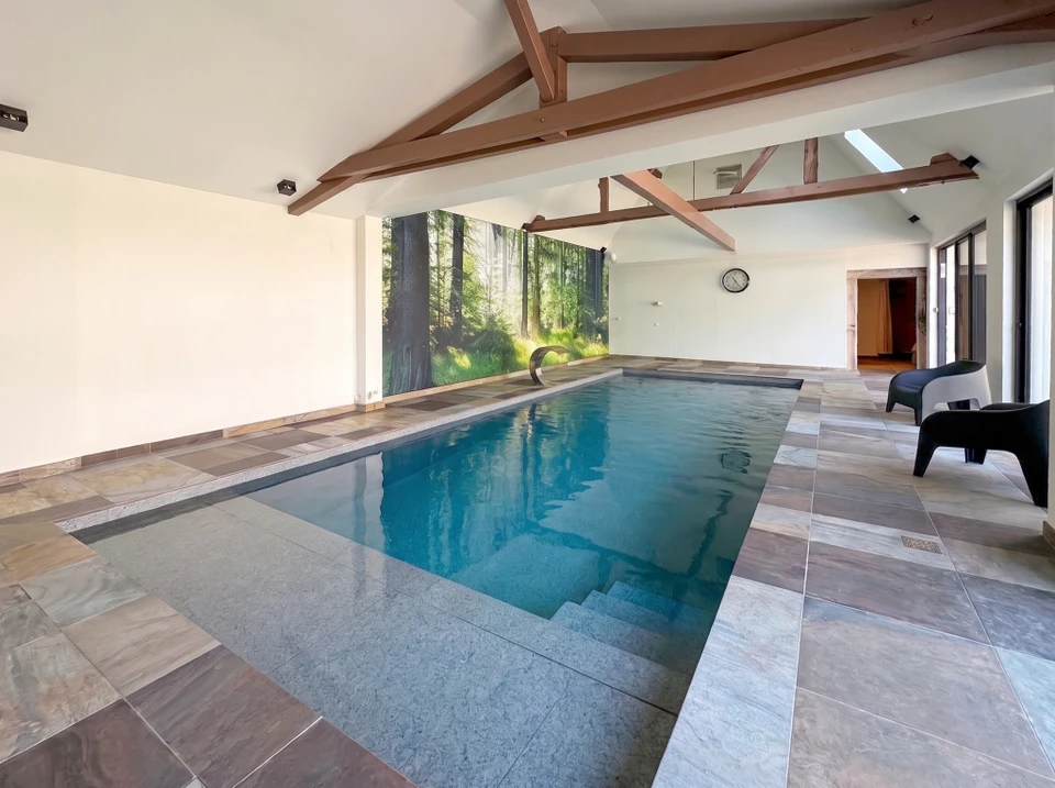 Piscine intérieure privée avec bassin rectangulaire carrelé bleu, escalier métallique avec garde-corps verre, plafond suspendu lamelles bois naturel, éclairage LED intégré spots encastrés, baies vitrées aluminium noir, sol carrelage grand format beige, appliques murales design triangulaires, architecture contemporaine haut de gamme