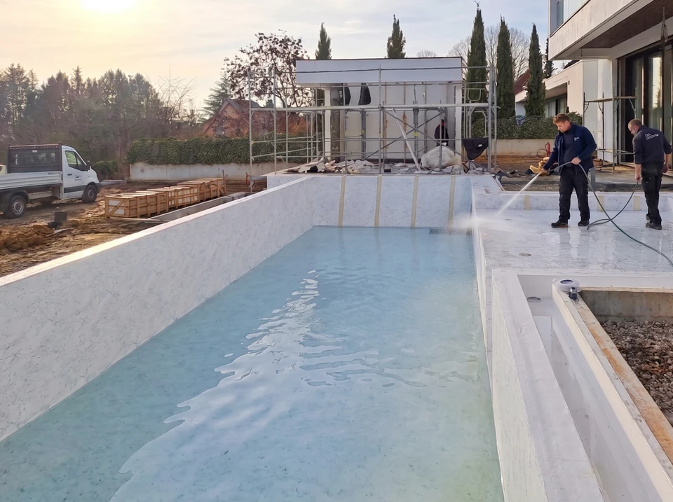 Piscine rectangulaire extérieure à débordement avec liner bleu ciel cristallin, entourée d'un dallage en grès cérame beige. Volet roulant immergé en aluminium anthracite côté arrière en position semi-ouverte. Pergola bioclimatique anthracite intégrée avec spa carré encastré en béton teinté visible. Panneau décoratif en acier corten oxydé avec oculus circulaire en arrière. Haies de bambous persistants et thuyas en clôture naturelle. Installation résidentielle haut de gamme, finition soignée, eau traitée limpide.